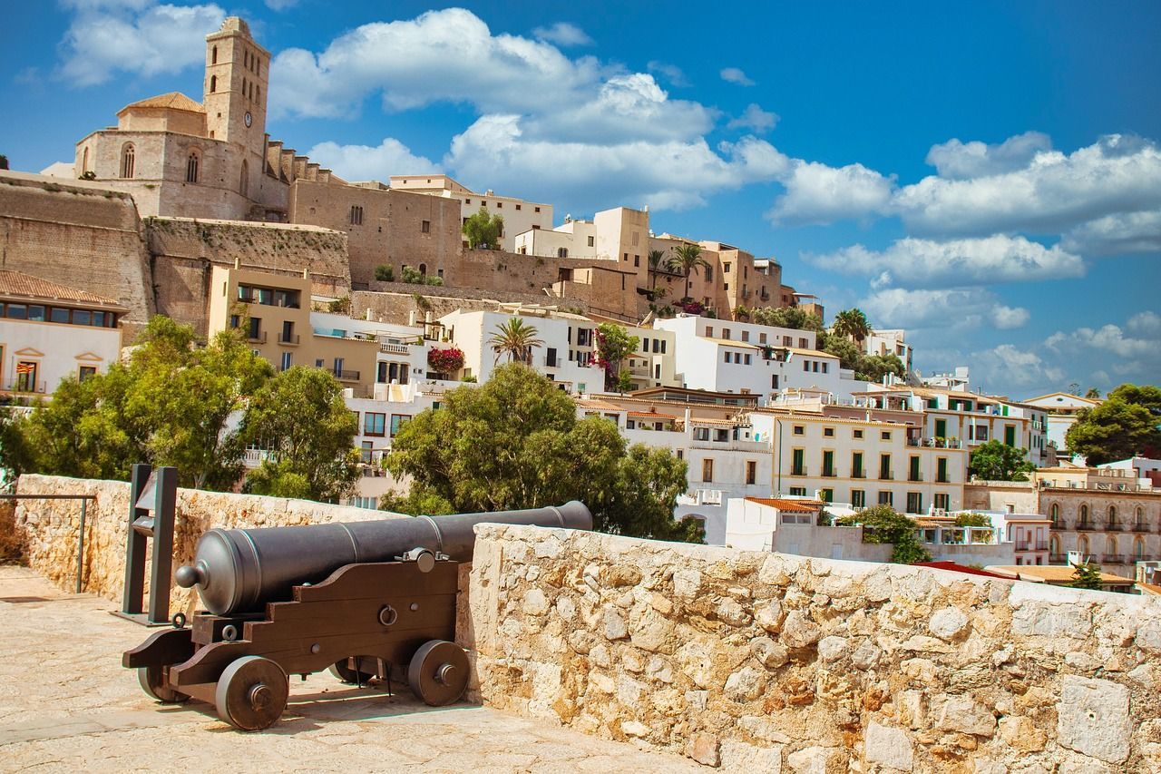 Un vieux canon repose sur un mur de pierre, dominant une ville historique à flanc de colline avec un clocher, sous un ciel bleu et des nuages blancs.