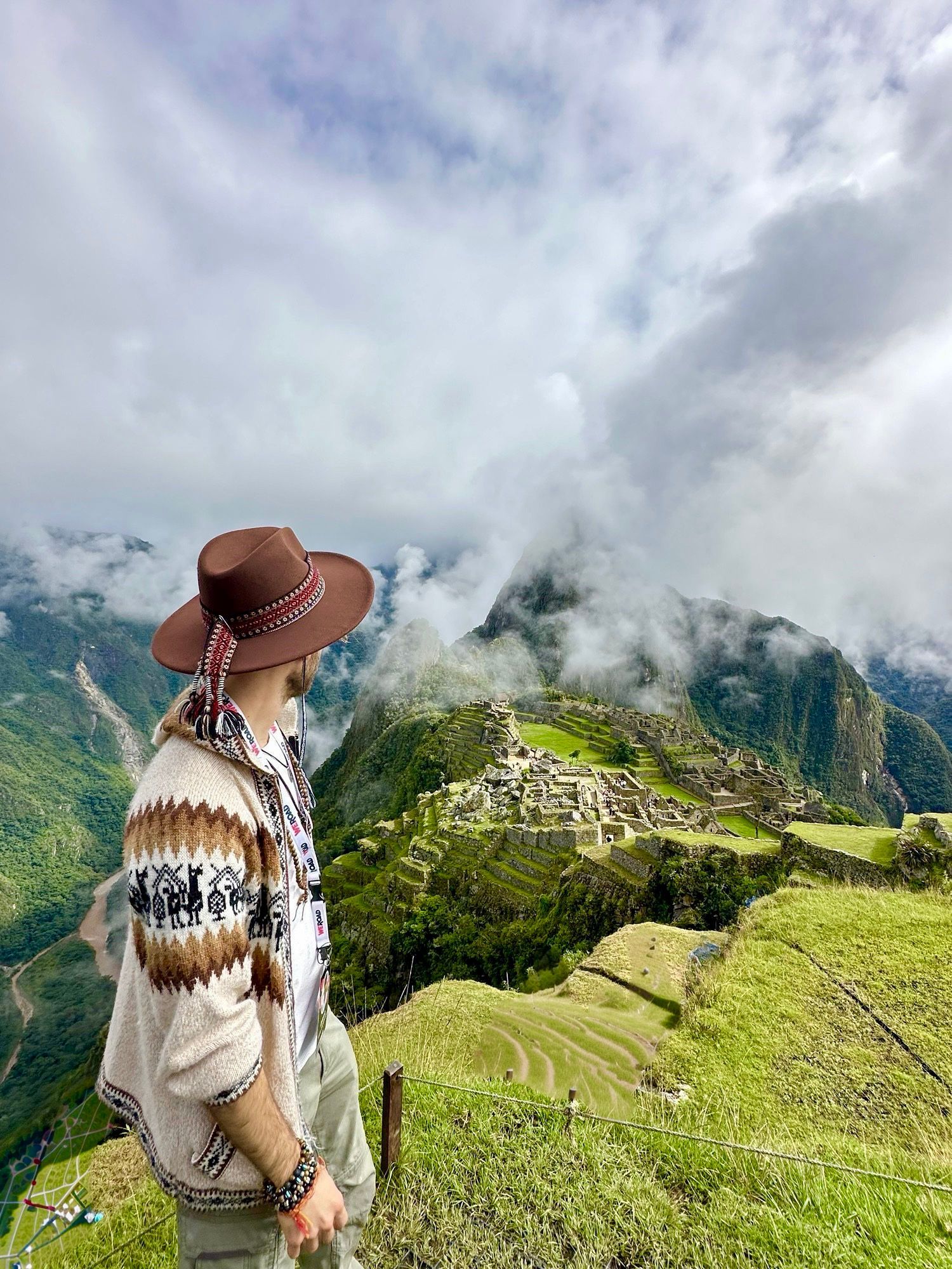 Una persona con un cardigan fantasia e un cappello marrone in piedi su una collina erbosa, con vista su antiche rovine immerse tra montagne verdi e nebbiose.