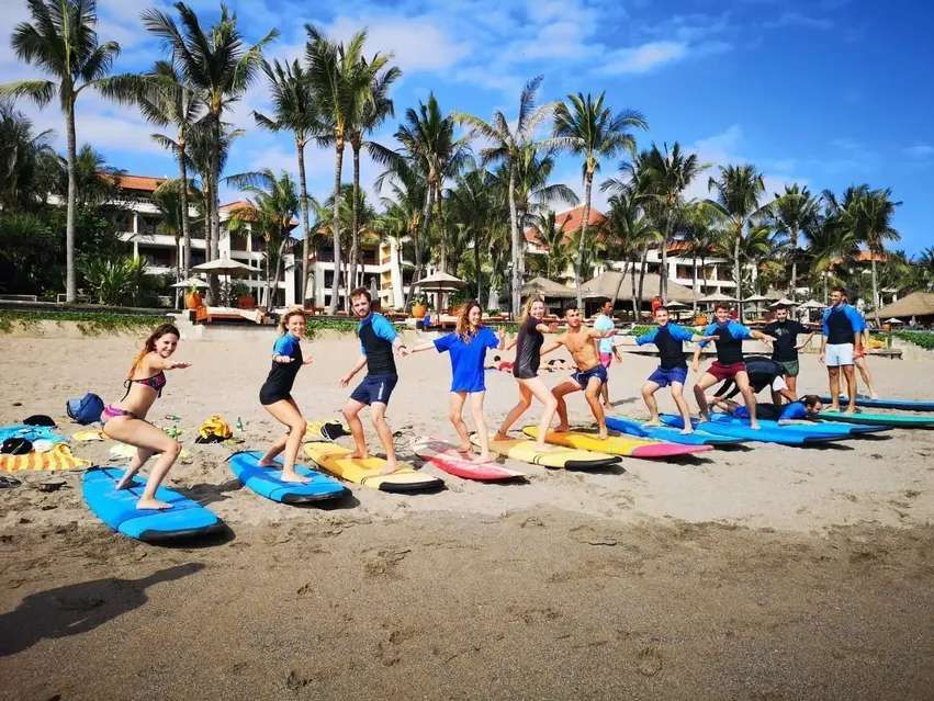 A WeRoad group trip lines up on a sandy beach for a surfing lesson, practicing their stances on colorful surfboards.