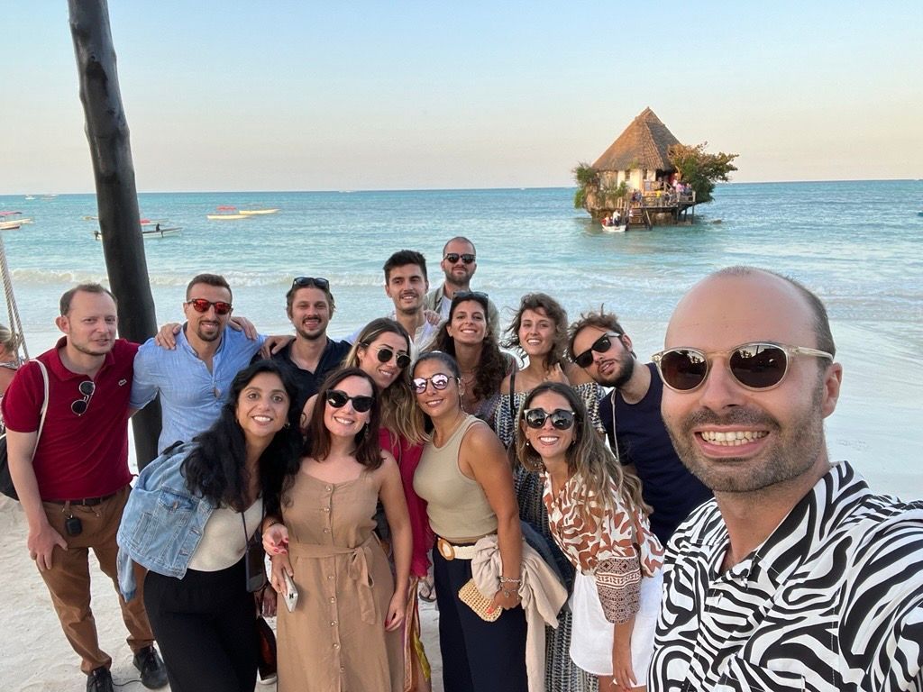 Un gruppo WeRoad si scatta un selfie sorridente su una spiaggia, con un ristorante nella roccia in mare sullo sfondo.