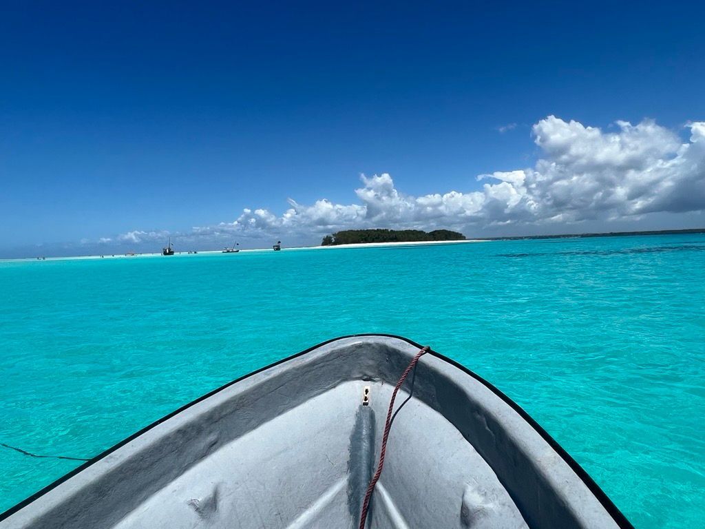 La prua di una barca naviga su acqua turchese verso un'isola lontana e alberata sotto un cielo blu con nuvole bianche.