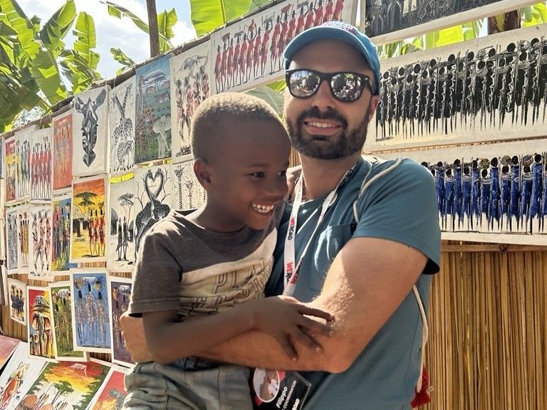 Un uomo di un viaggio di gruppo WeRoad sorride mentre tiene in braccio un bambino davanti a una bancarella d'arte all'aperto.