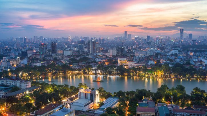 An aerial view of a sprawling cityscape with an illuminated lake at dusk, under a pink and blue sky.