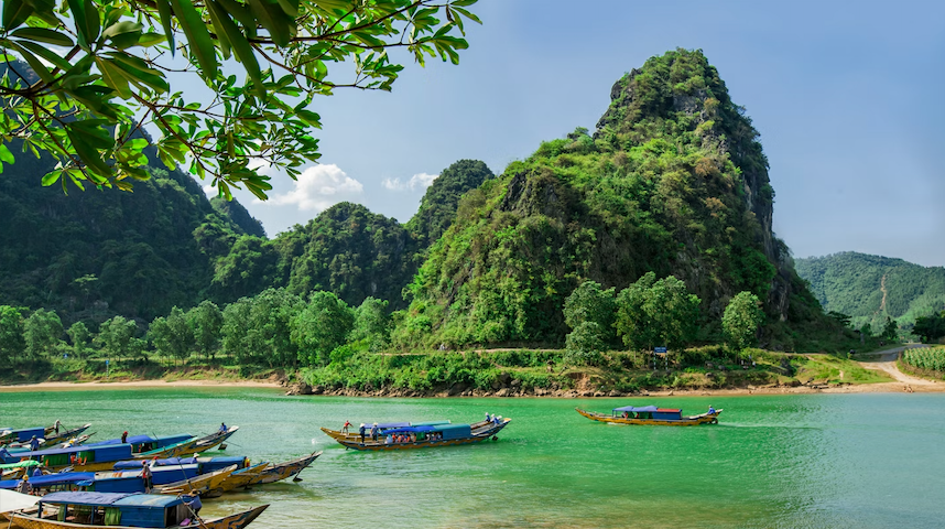 Traditional long boats on a vibrant green river at the base of lush, forest-covered mountains under a sunny sky.