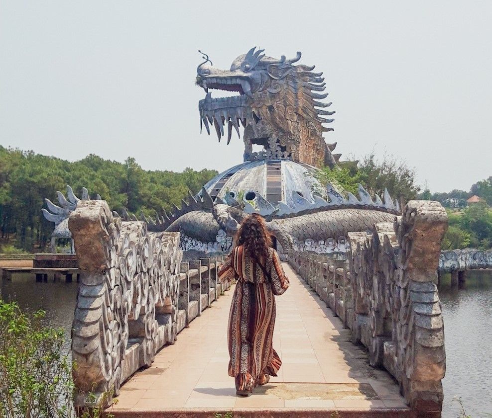 A woman seen from behind walks on a stone bridge towards a large, weathered sculpture of a dragon built over a lake.