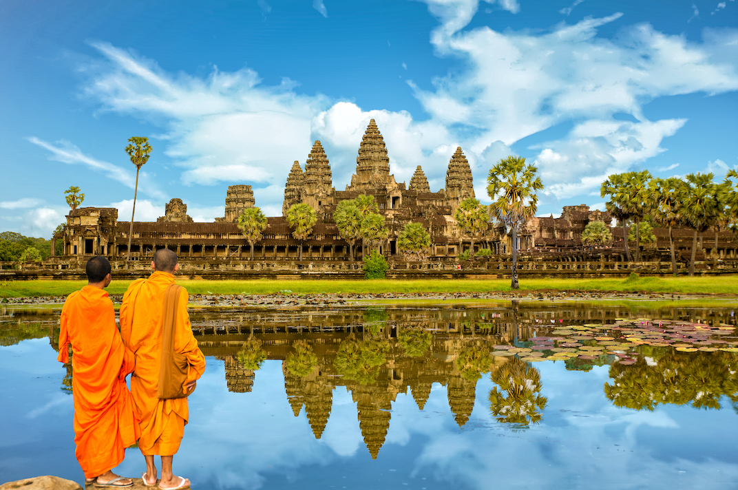 Two monks in orange robes stand by a lake, looking at an ancient stone temple reflected in the calm water.