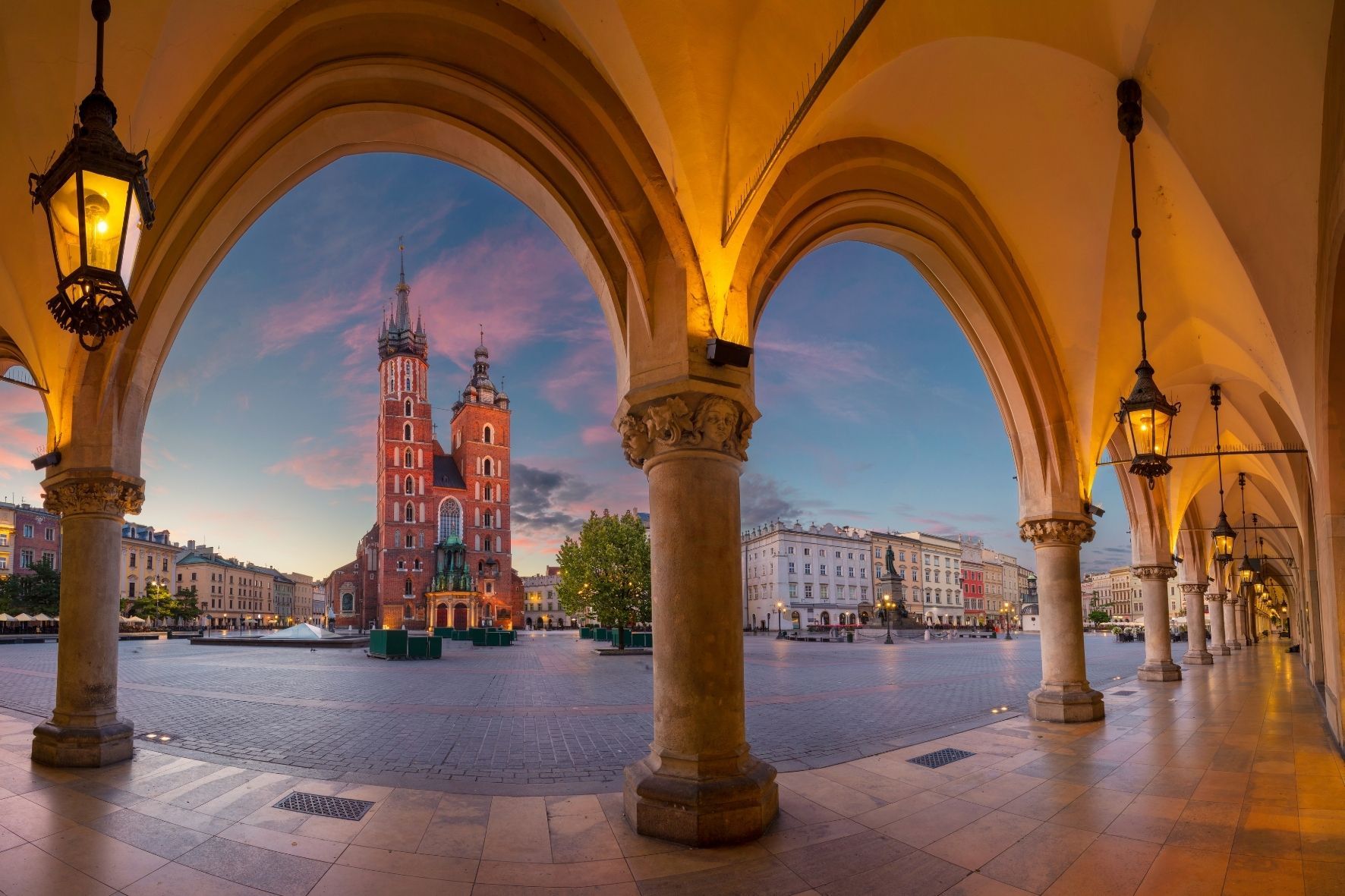 Una iglesia histórica de ladrillo rojo con dos torres se erige en una plaza de la ciudad, observada desde debajo de arcos de piedra iluminados al atardecer.