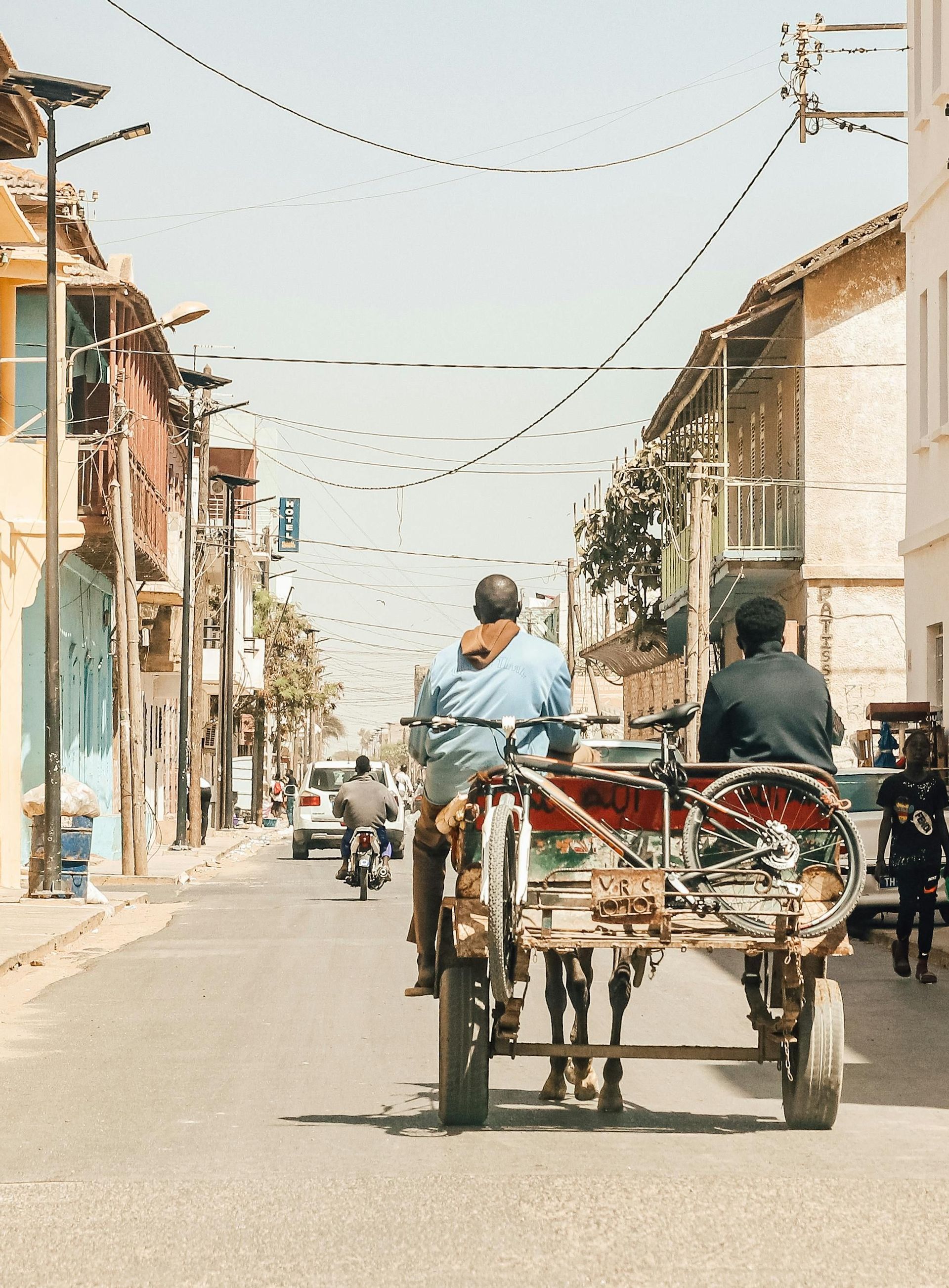 Vista trasera de dos hombres en un carro de caballos con una bicicleta adjunta, por una calle soleada con edificios.