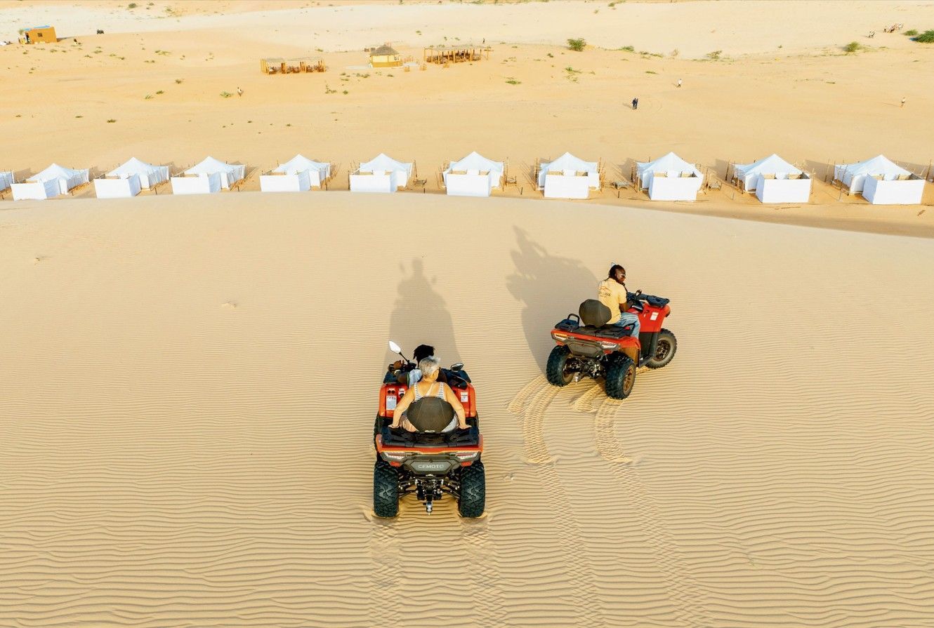 Vista aérea de dos personas de un viaje en grupo de WeRoad en quads por dunas de arena, con un campamento en el desierto visible al fondo.