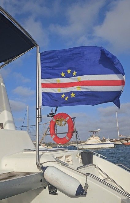 La bandera de Cabo Verde ondeando al viento, montada en la popa de un barco en un puerto deportivo bajo un cielo azul.