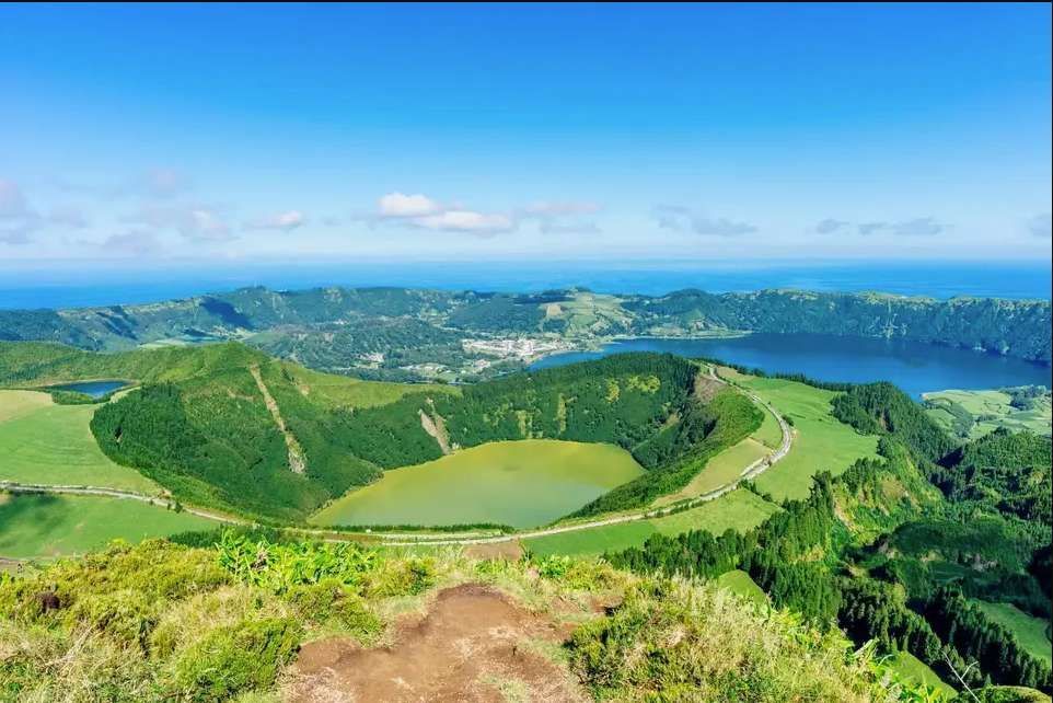 A panoramic view of two volcanic crater lakes, one green and one blue, nestled among lush hills with the ocean in the distance.