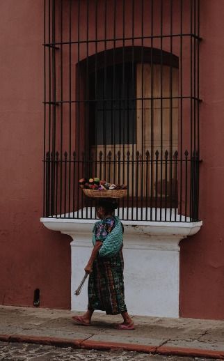A woman in traditional clothing walks on a sidewalk, balancing a woven basket of produce on her head next to a reddish-brown wall.
