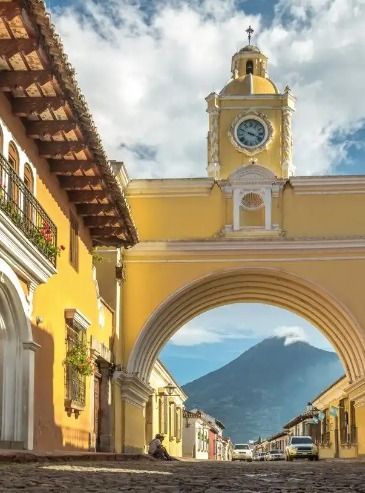 Un arco giallo con una torre dell'orologio si staglia su una strada di ciottoli, incorniciando la vista di un grande vulcano in lontananza sotto un cielo azzurro.