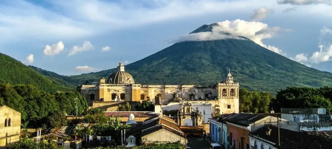 Una città coloniale con una grande chiesa a cupola si trova ai piedi di un vulcano verdeggiante, con la cima avvolta dalle nuvole, sotto un cielo azzurro.