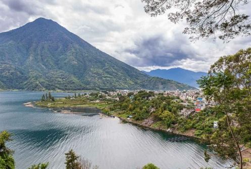 Una città lacustre si trova su una penisola ai piedi di una grande montagna boscosa sotto un cielo nuvoloso.
