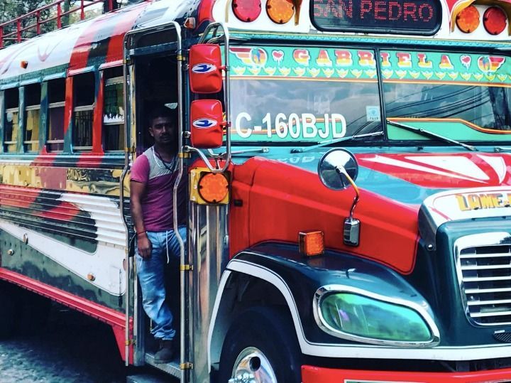 Un uomo con una maglietta rosa e jeans è in piedi sulla porta aperta di un autobus dai colori vivaci e con dettagli cromati.