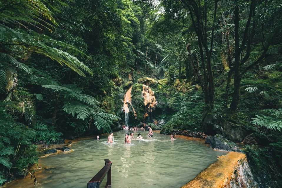 A WeRoad group trip swimming in a natural thermal pool with a waterfall, surrounded by a dense green forest.