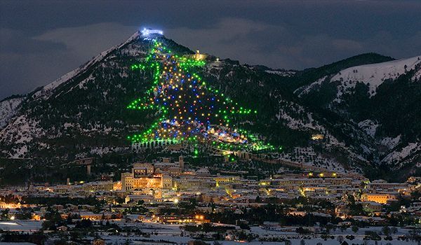 Un immenso albero di Natale fatto di luci colorate illumina una montagna innevata sopra un paese illuminato di notte