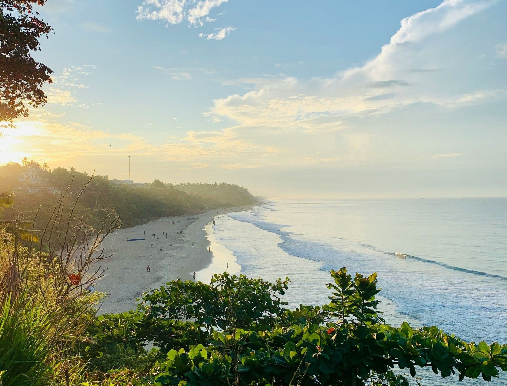 Vue aérienne d'une longue plage de sable lors d'un lever de soleil doré, avec des personnes sur le rivage et des vagues océaniques calmes.