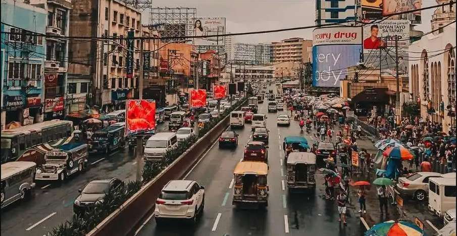 An elevated view of a busy city street with heavy traffic on a rainy day, surrounded by buildings and billboards.