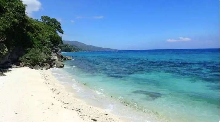 A white sand beach meets clear turquoise water next to a rocky, tree-covered shoreline under a blue sky.