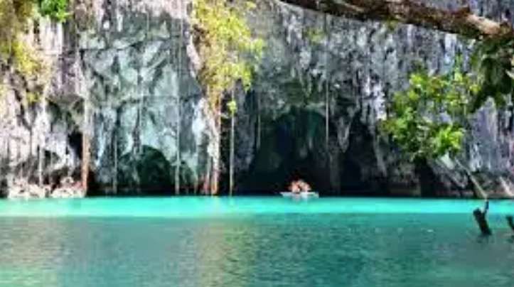 A small boat with people floats on turquoise water at the entrance of a large sea cave with rocky cliffs and hanging vines.