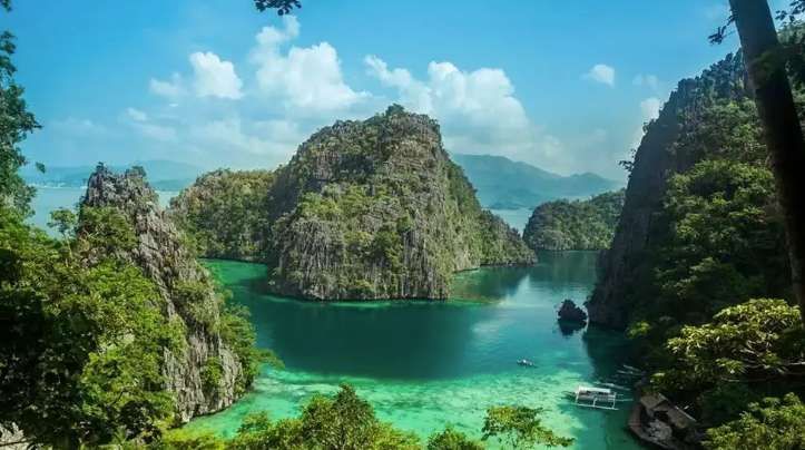 A high-angle view of a tropical lagoon where lush, green-covered limestone cliffs rise from clear turquoise water under a partly cloudy sky.