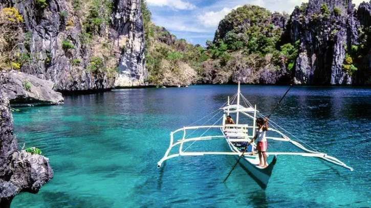 A person navigates a traditional outrigger boat across a turquoise lagoon, enclosed by steep, jungle-covered rock formations.