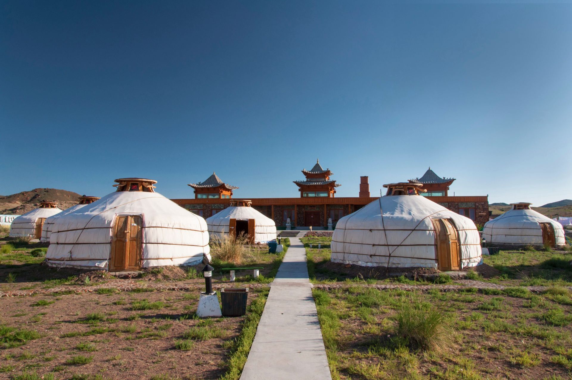 Plusieurs yourtes blanches sont installées dans un champ verdoyant, devant un grand bâtiment de style traditionnel, sous un ciel bleu dégagé.