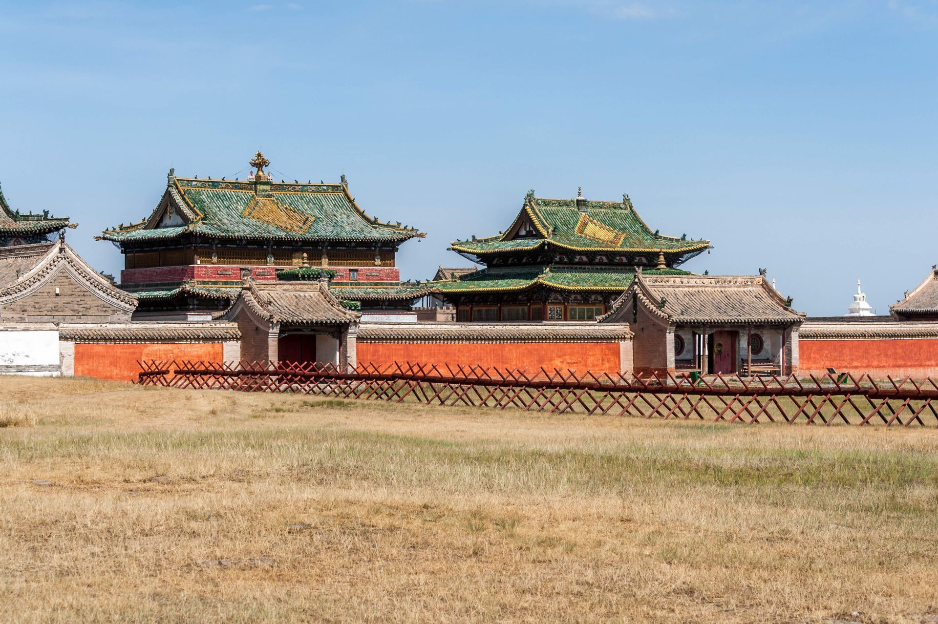 A Mongolian monastery with green tiled roofs and a red perimeter wall, viewed from across a dry grassy field under a clear blue sky.