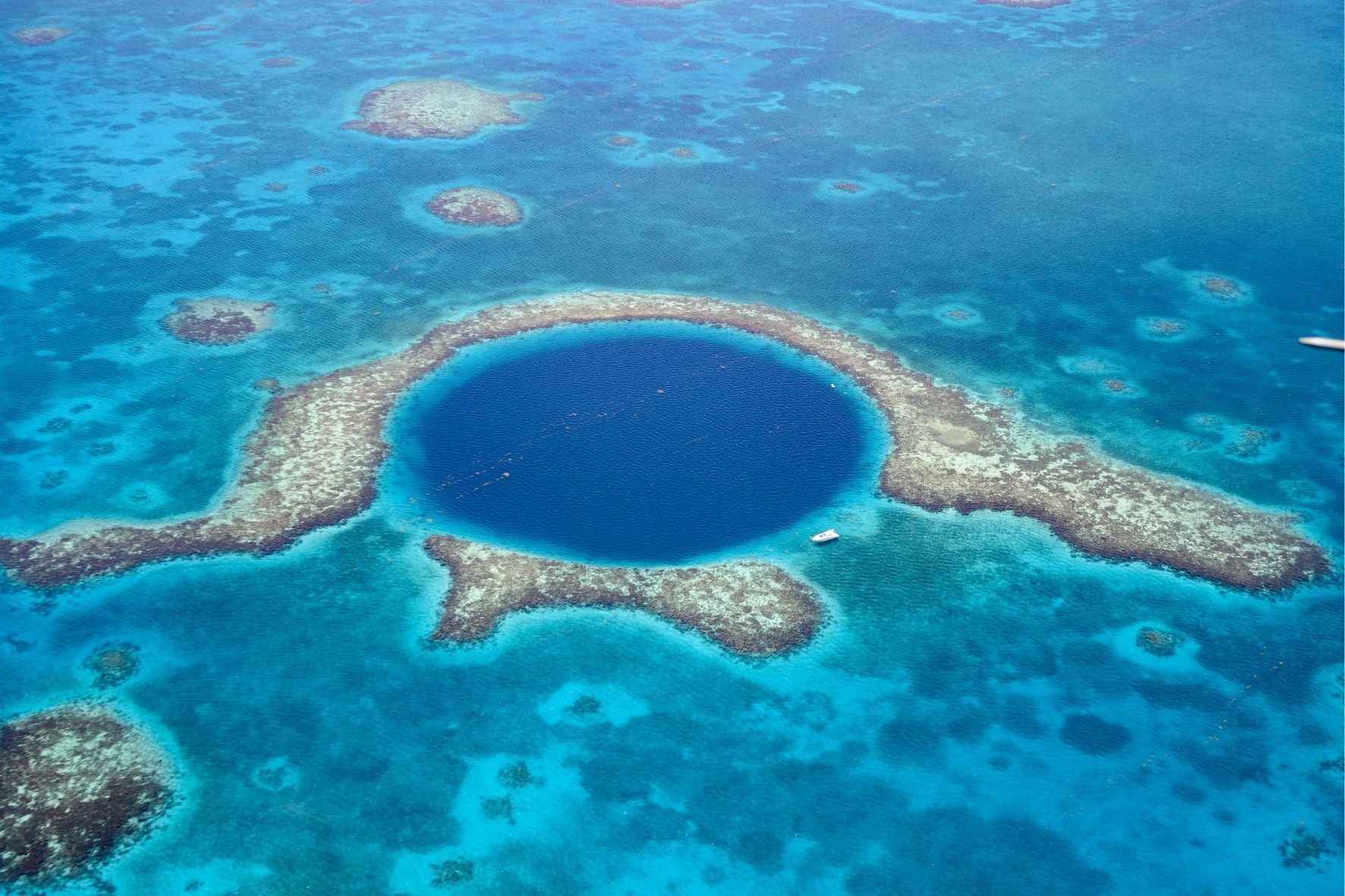 An aerial view of a large, deep blue marine sinkhole surrounded by a coral reef in a vast turquoise ocean.