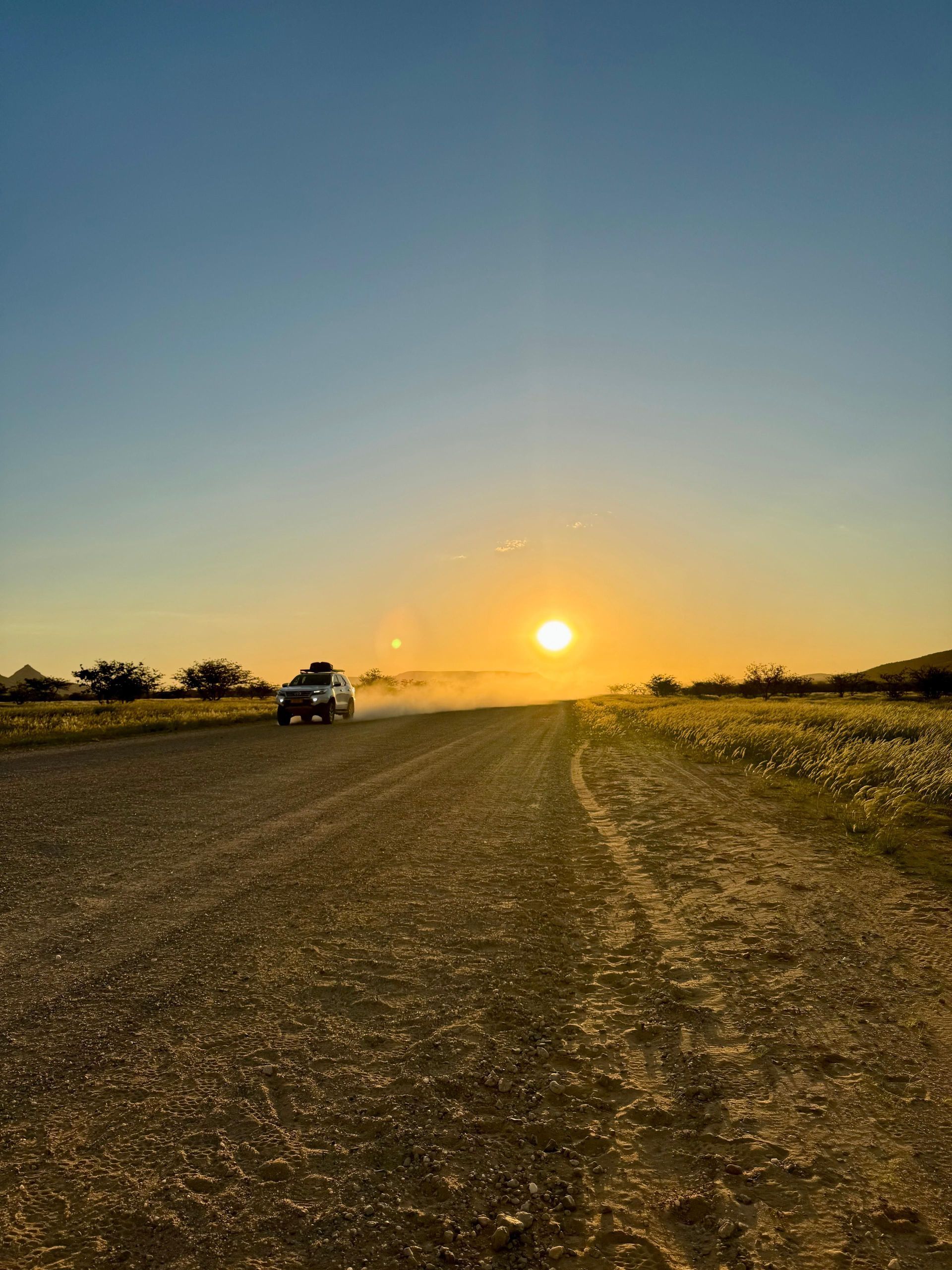 Un SUV bianco con bagagli sul tetto percorre una strada sterrata polverosa, sollevando polvere contro un tramonto dorato.