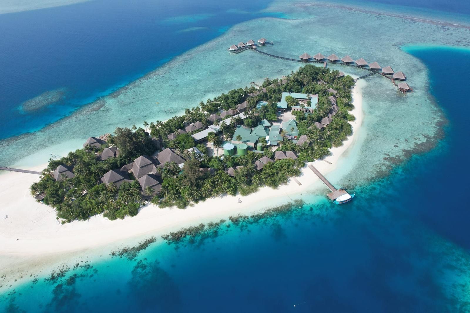 Una vista aérea de un resort en una isla tropical con playas de arena blanca, palmeras y bungalows sobre el agua rodeados por el océano turquesa.
