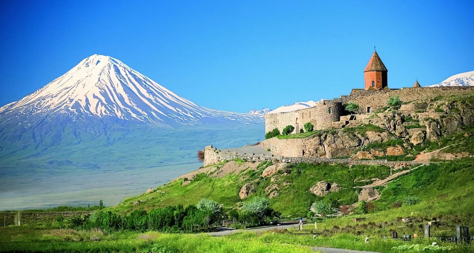 Eine steinerne Festung liegt auf einem grasbewachsenen, felsigen Hügel, mit einem großen, schneebedeckten Berg im Hintergrund vor einem klaren blauen Himmel.