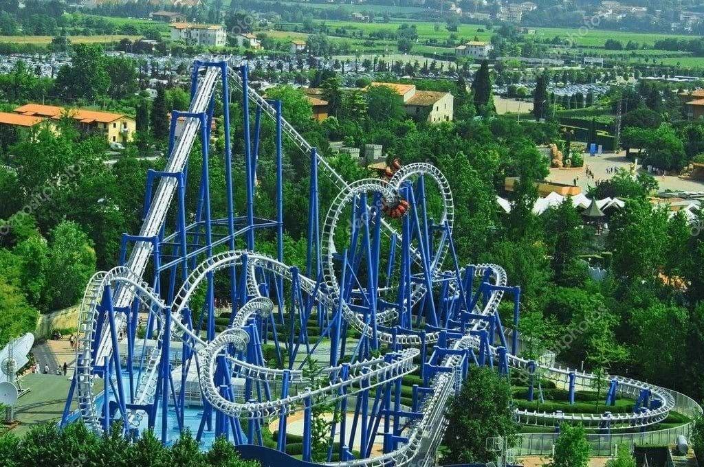 Vista aerea di imponenti montagne russe blu e bianche, con numerosi anelli e avvitamenti, immerse nel verde lussureggiante di un parco divertimenti.