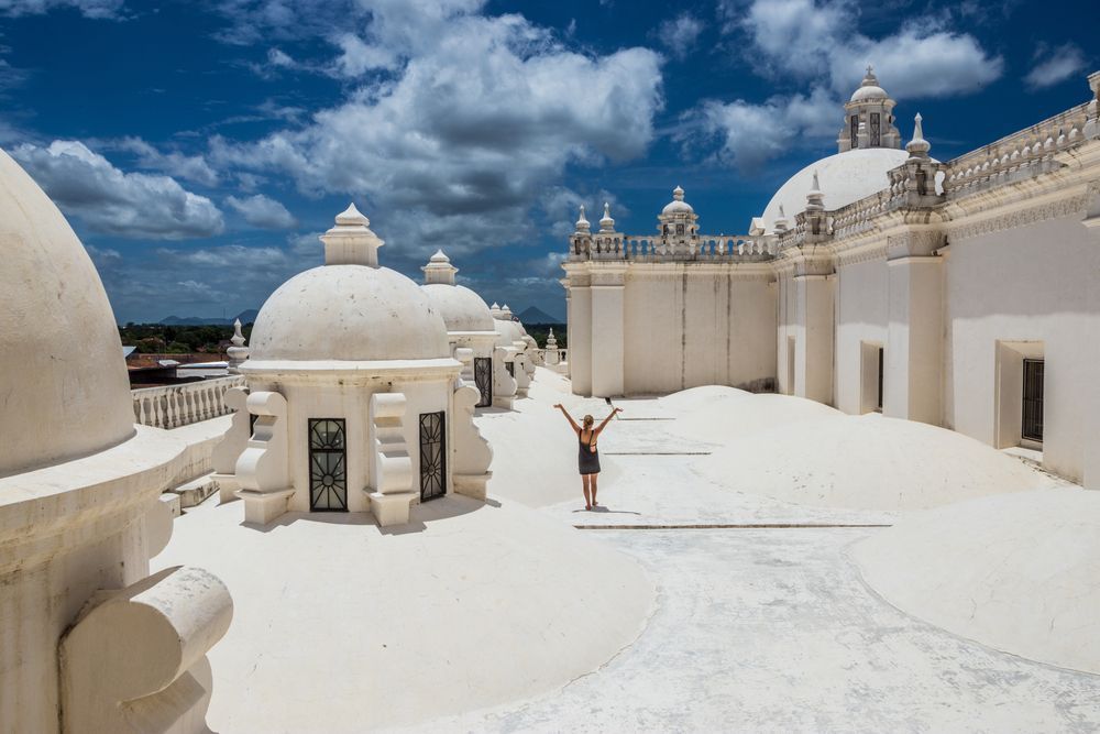 Una donna con le braccia alzate è in piedi sul vasto tetto bianco di un edificio con cupole multiple sotto un cielo blu nuvoloso.