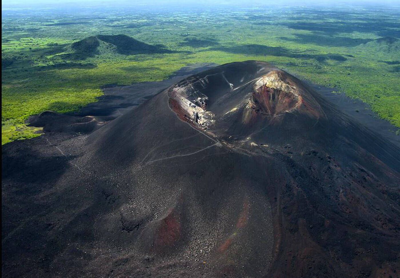 Una vista aerea di un vulcano scuro con escursionisti sul pendio che si dirigono verso il cratere, immerso in un lussureggiante paesaggio verde.