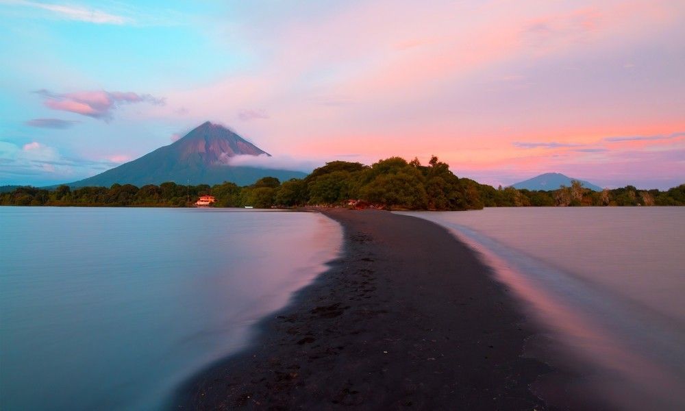Una lingua di sabbia nera si curva in un lago calmo, con un grande vulcano visibile sullo sfondo sotto un cielo colorato al tramonto.