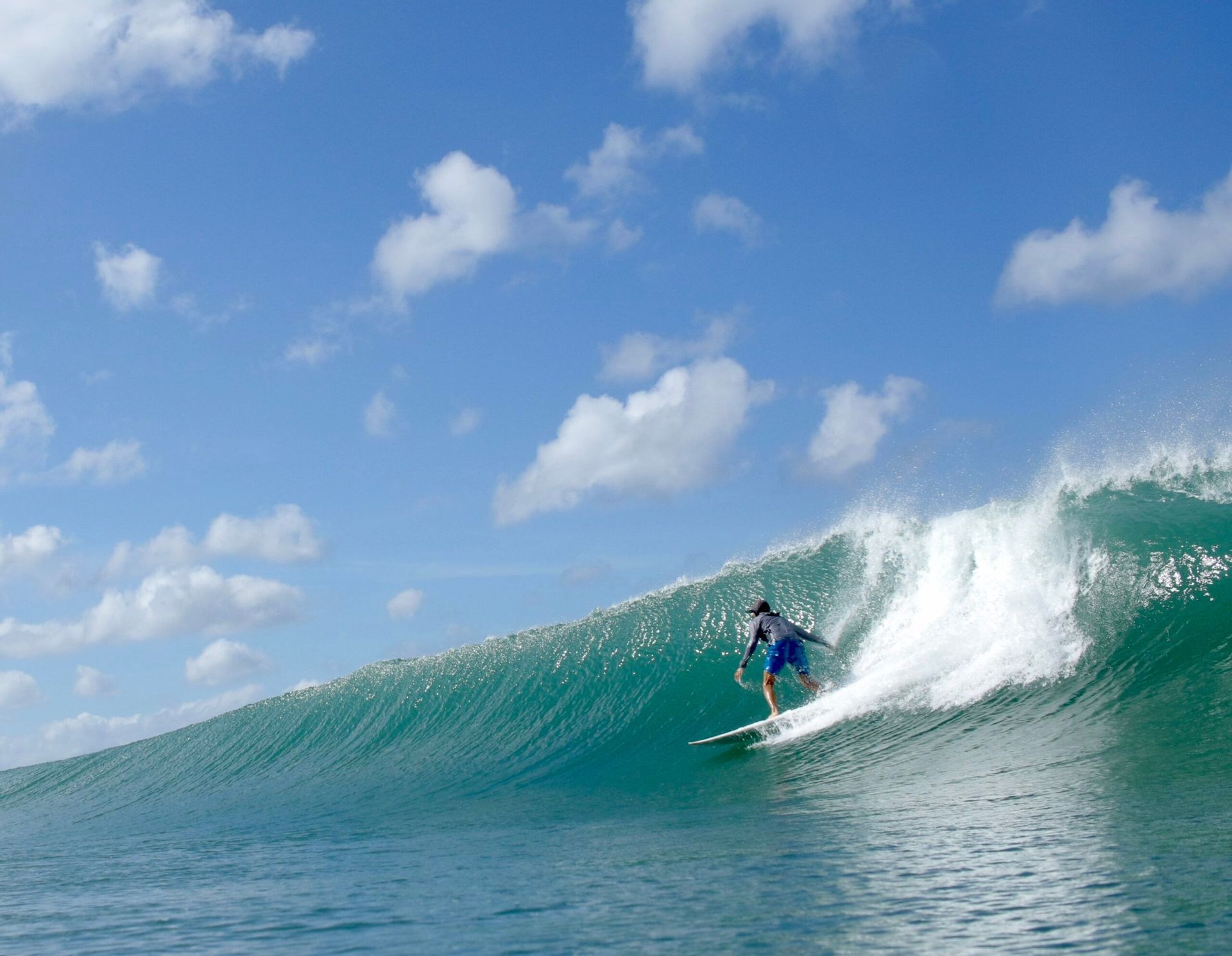 Un surfista in pantaloncini blu cavalca una grande onda turchese sotto un cielo parzialmente nuvoloso.