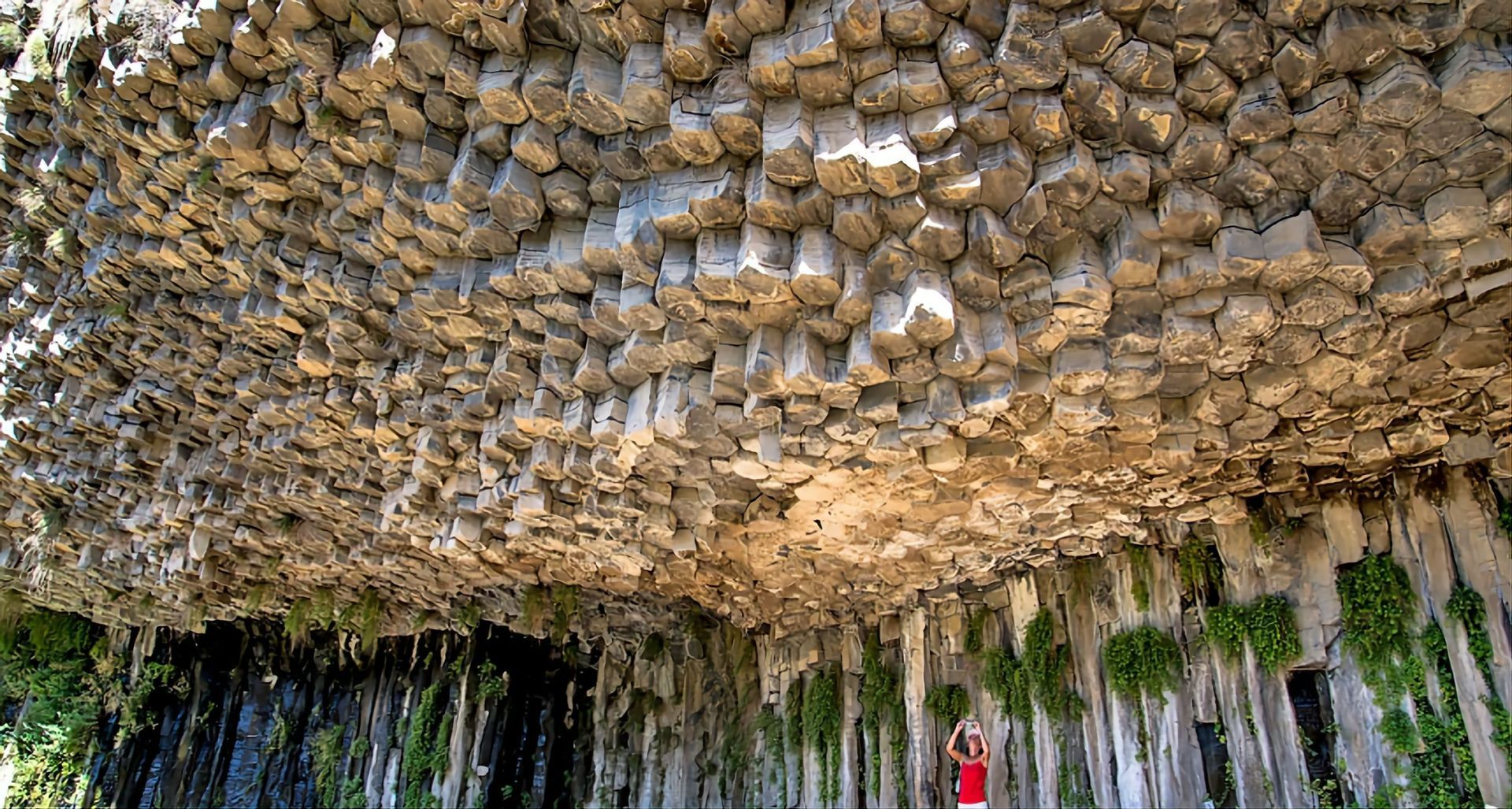 Eine Person in einem roten Oberteil steht und blickt auf eine riesige Decke aus sechseckigen Basaltsäulen in einer Felshöhle.