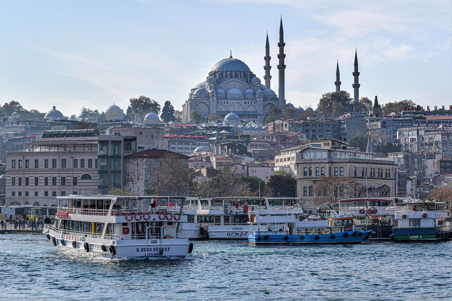 Ferries sail on the water before a dense cityscape, with a large mosque featuring a central dome and tall minarets on a hill.