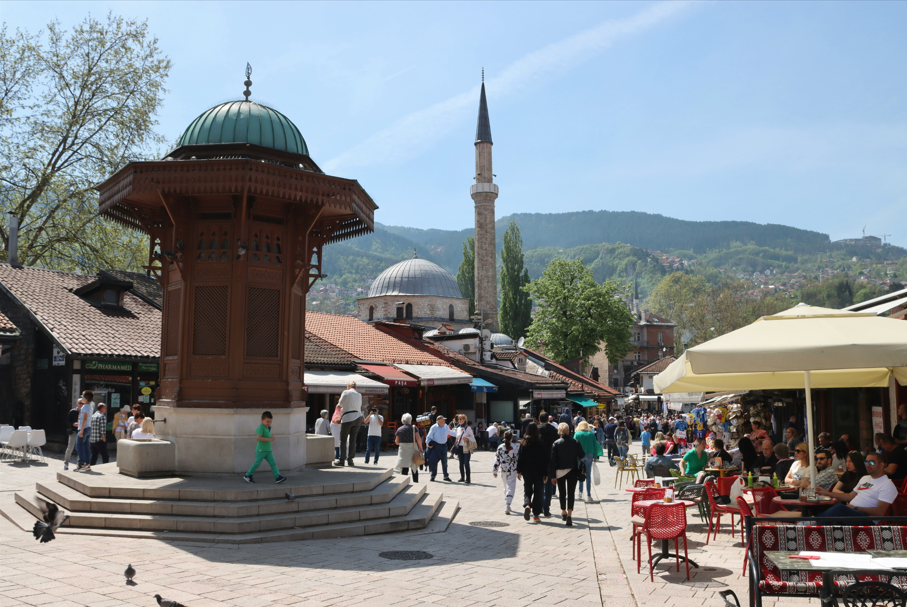 Une grande fontaine en bois se dresse sur une place de ville animée, avec une mosquée en arrière-plan et des gens se promenant parmi les cafés en plein air.