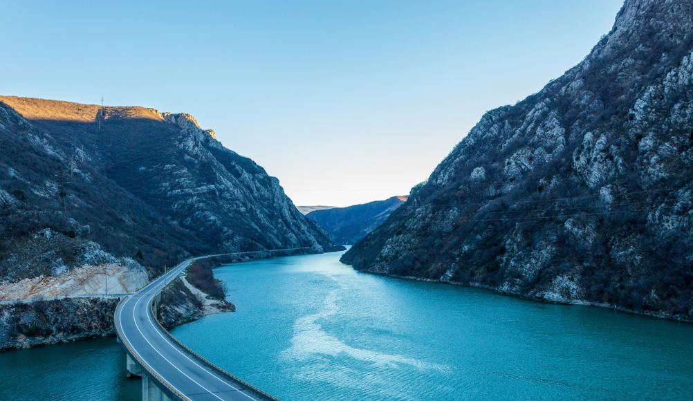 Vue aérienne d'un pont avec une route sinueuse longeant une rivière bleu vif traversant un canyon montagneux escarpé.