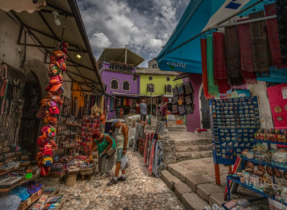 Les gens flânent devant les étals de souvenirs et de textiles d'un marché en plein air, dans une rue pavée étroite, entre des bâtiments colorés.