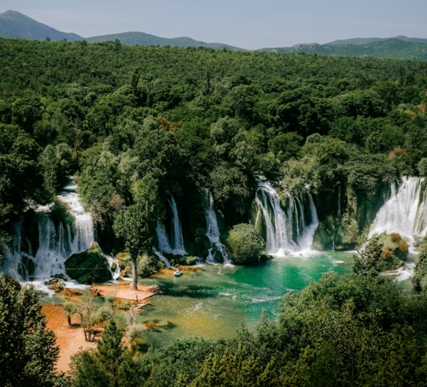 Vue panoramique de multiples cascades se déversant dans un bassin turquoise, entouré d'une forêt verte dense avec des collines en arrière-plan.