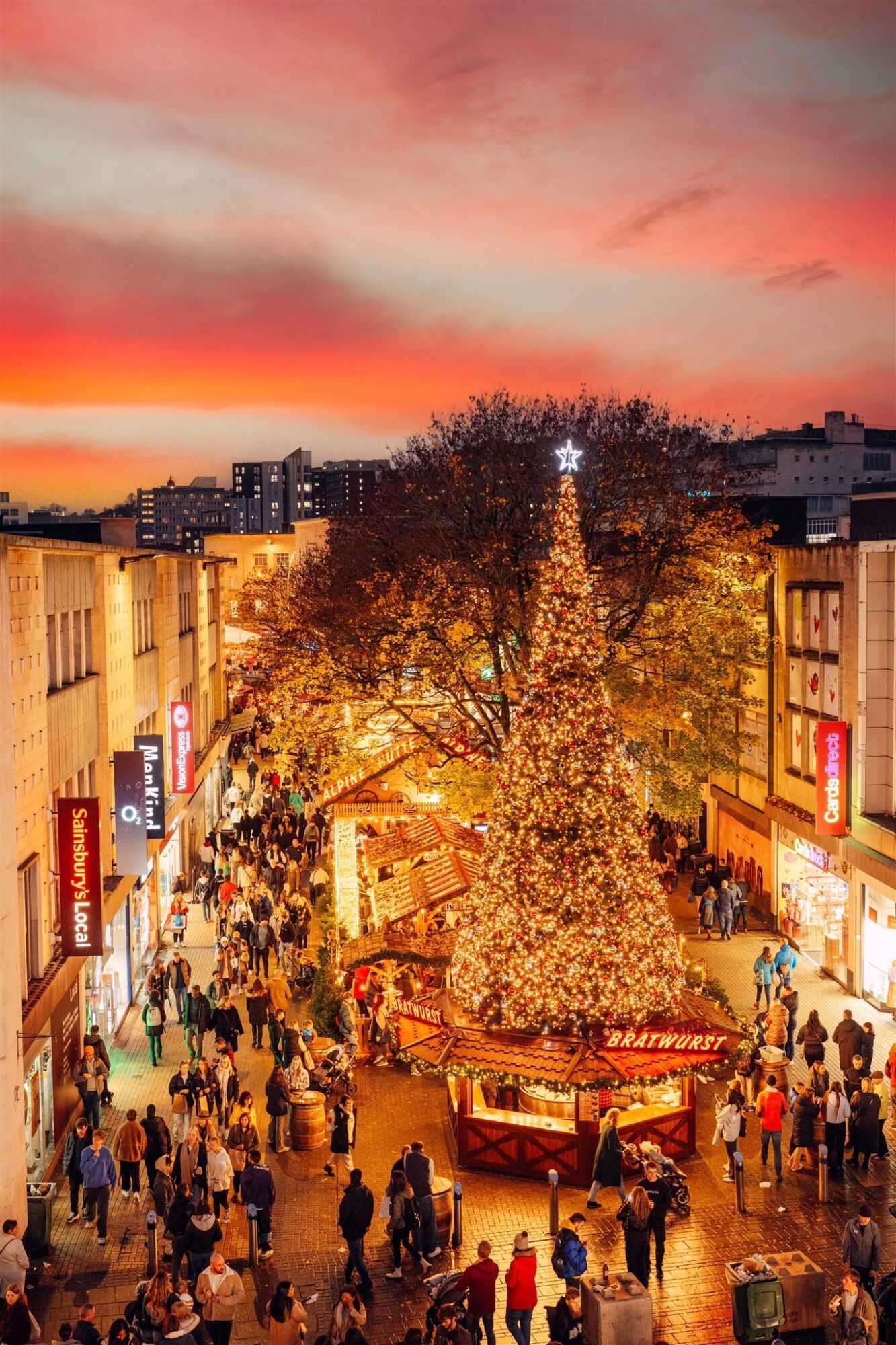 Una veduta dall'alto di una vivace strada cittadina con un mercatino di Natale, caratterizzata da un grande albero di Natale illuminato e folle sotto un cielo al tramonto vibrante.