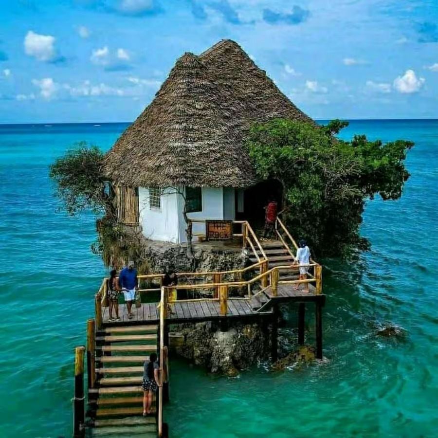 Une maison au toit de chaume construite sur un rocher dans l'océan turquoise, avec des gens marchant sur un escalier en bois menant à l'entrée.