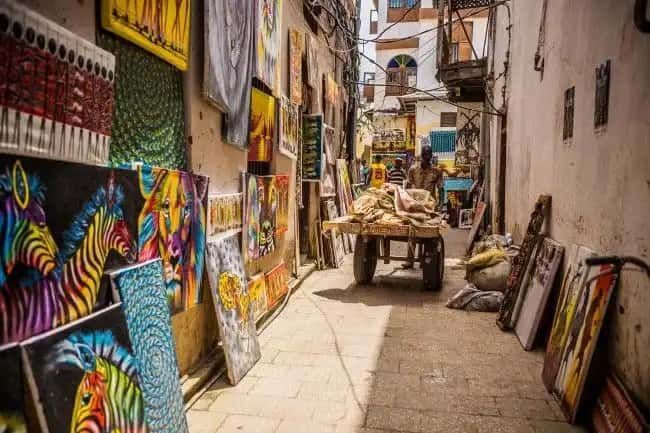 Un marché dans une ruelle étroite et ensoleillée, avec des peintures colorées de zèbres et d'autres animaux exposées sur les murs à vendre.