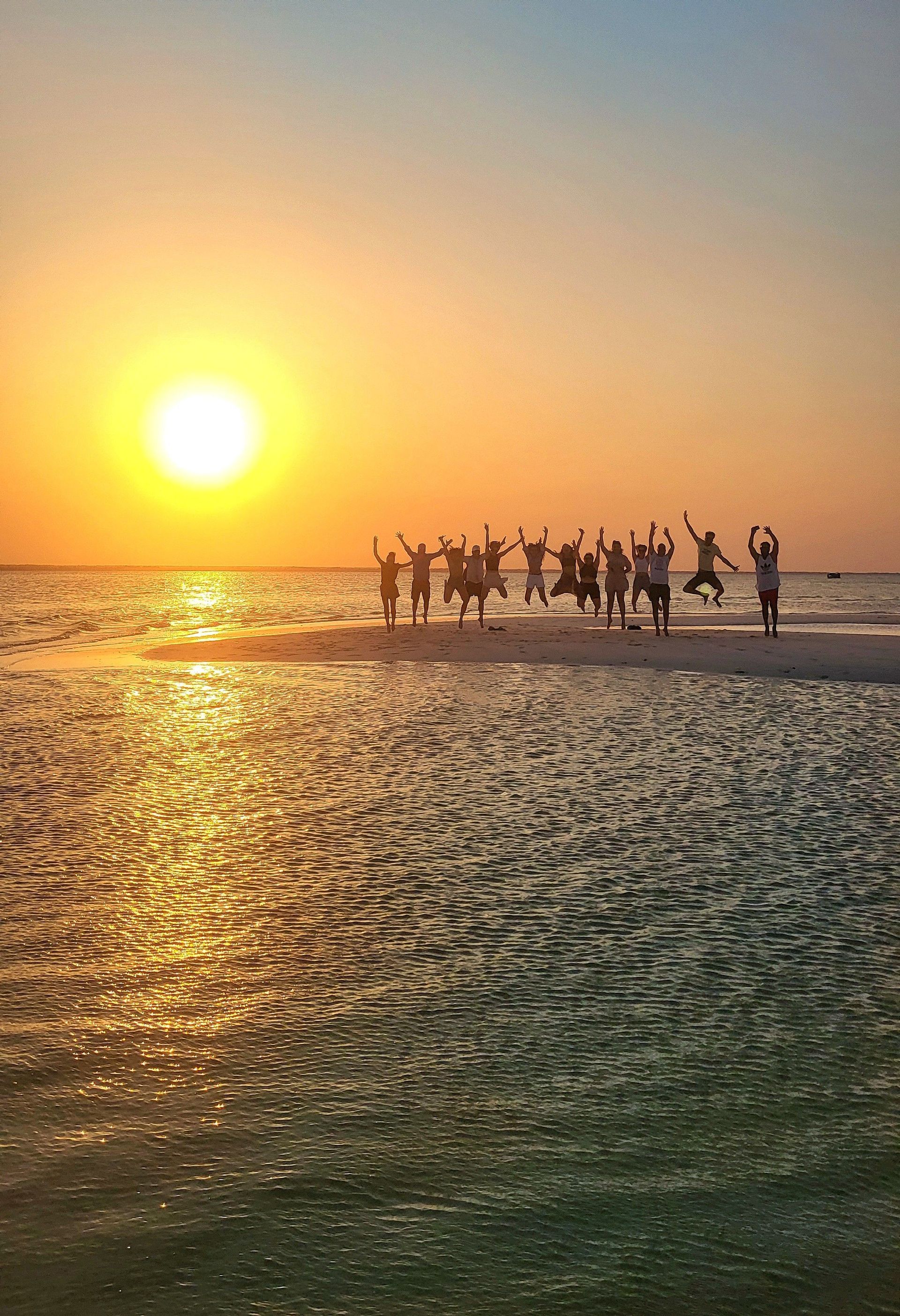 Eine WeRoad-Gruppe springt als Silhouette auf einer Sandbank, während die Sonne über dem ruhigen Meer versinkt.