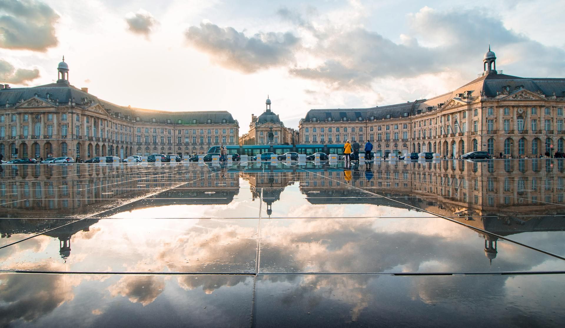 Un grande edificio classico e un cielo nuvoloso si riflettono nel terreno bagnato di una grande piazza pubblica al tramonto.